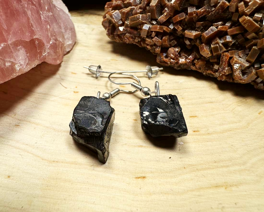 A pair of raw elite noble shungite earrings displayed on a rustic wooden surface with decorative crystals in the background.