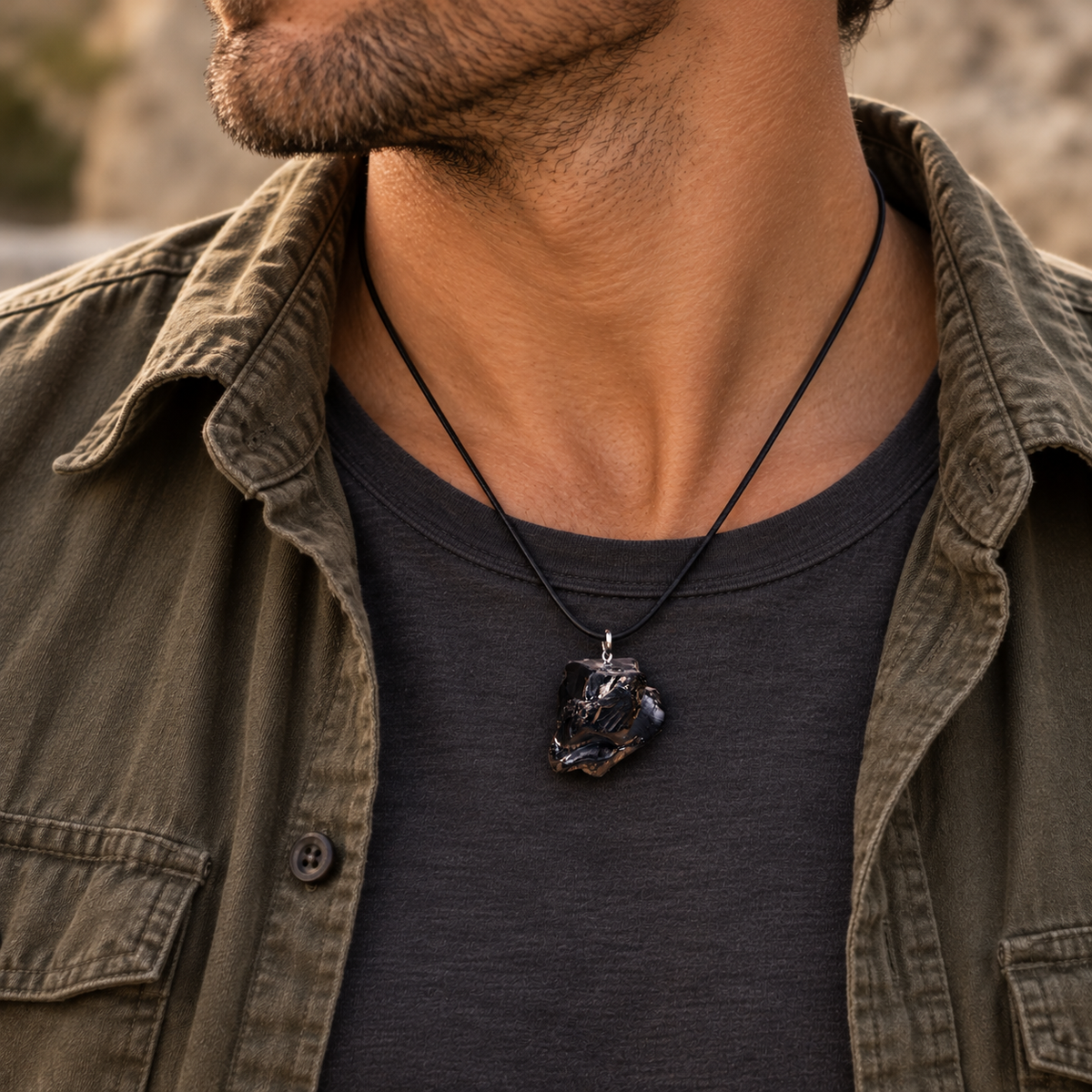 Close-up of a man wearing a raw metallic black Elite Noble Shungite stone pendant necklace over a grey t-shirt and an open olive green button-up shirt.