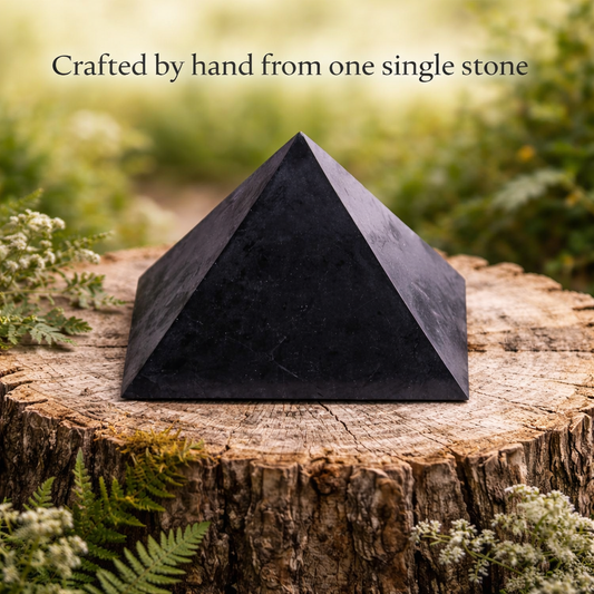 A large polished black shungite pyramid displayed outdoors on a rustic tree stump with overlay text reading 'Crafted by hand from one single stone'.