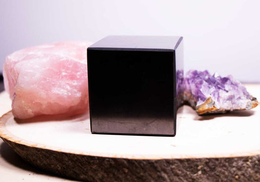 A polished black authentic shungite cube sitting on a natural wood slice tray, flanked by a raw pink rose quartz block and an amethyst crystal cluster.