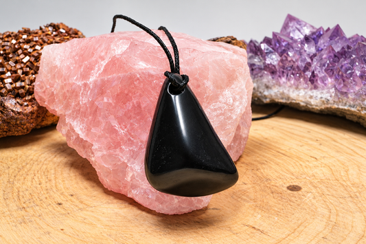 A glossy, polished black tumbled Shungite Harmony Pendant necklace draped directly over a large specimen of raw pink rose quartz. The background shows a cluster of purple amethyst crystals arranged on a natural wood surface.