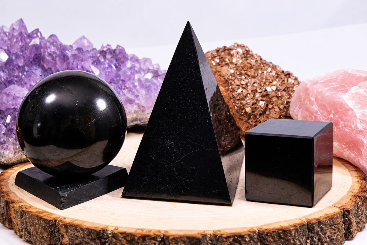 An assortment of polished black authentic shungite shapes, including a sphere on a stand, a Golod pyramid, and a geometric cube, displayed on a natural wood slice tray with raw crystal clusters in the background.