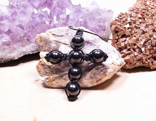 An authentic black Shungite cross pendant handcrafted from six individually carved stone beads, displayed on a natural matrix rock next to a purple amethyst crystal.