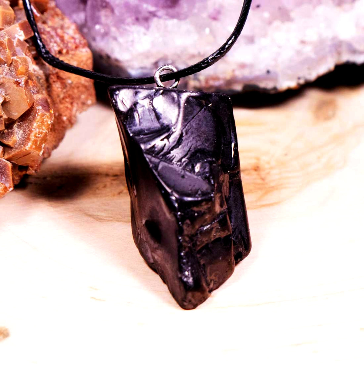 Raw metallic black Elite Noble Shungite stone pendant attached to a black cord necklace, displayed on a natural light wood background next to amethyst crystals.