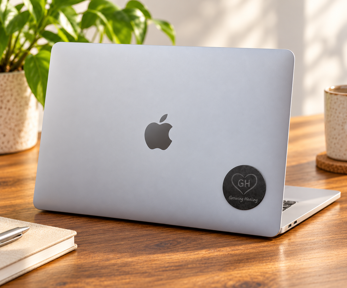 A black circular Shungite tech tab featuring a heart logo attached to the bottom right corner on the back of a silver MacBook laptop.