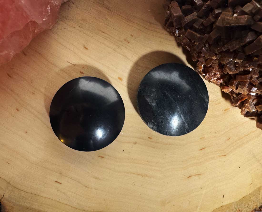 An alternate close-up view of a pair of round, polished black shungite pocket polarity harmonizer stones on a natural wood slice.
