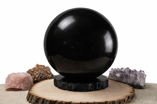 A large polished black stone sphere resting on a dark circular base over a natural wood slice stand, arranged with a raw pink quartz piece and a purple amethyst cluster.