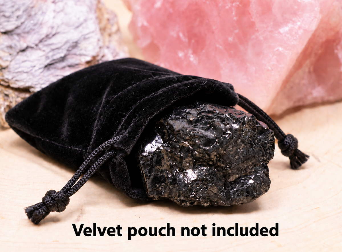 A large, raw elite noble shungite crystal with a metallic silver sheen resting in a black velvet drawstring bag on a wooden base with overlay text reading 'Velvet pouch not included'