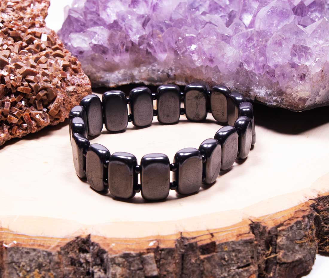 Straight-on view of a men's black Shungite grounding bracelet with polished rectangular tiles, set against a natural raw cluster and purple amethyst crystal background.