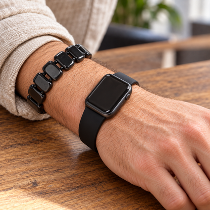 Close-up lifestyle view of a man's wrist wearing an authentic black Shungite grounding bracelet next to a space gray Apple Watch on a wooden desk.