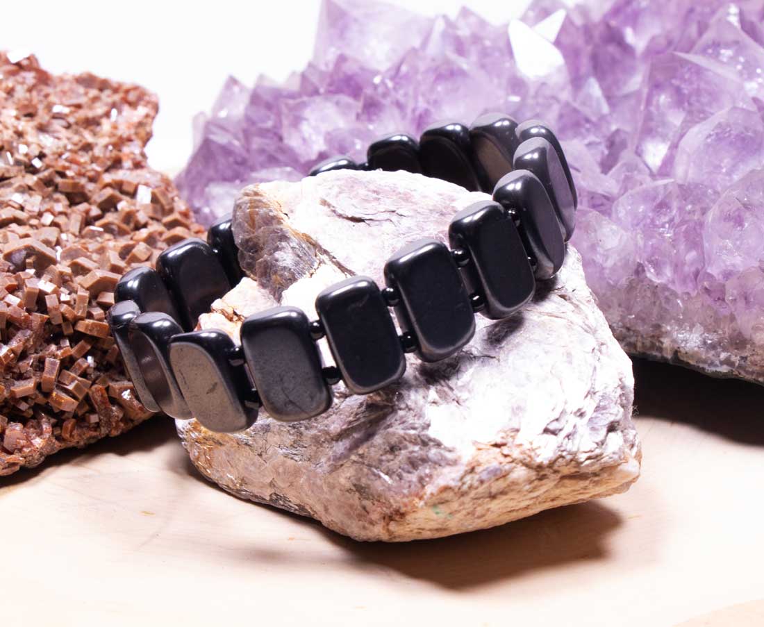 Men's black Shungite grounding bracelet with flat rectangular beads displayed on natural stones with purple amethyst crystal background.