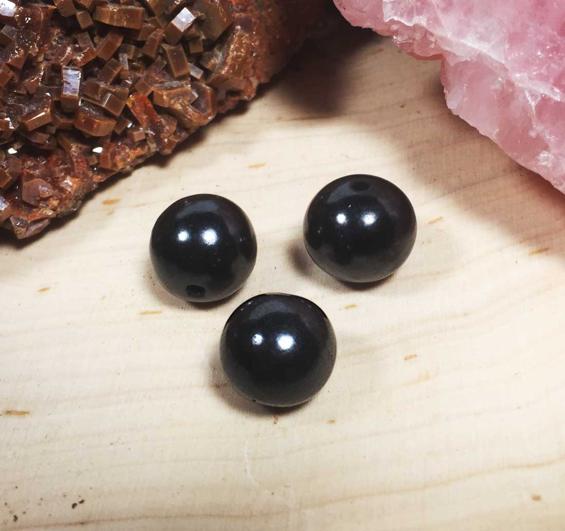 A set of three loose spherical Shungite stone beads for jewelry making, displayed with rustic crystals in the blurred background.
