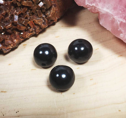 A set of three loose spherical Shungite stone beads for jewelry making, displayed with rustic crystals in the blurred background.
