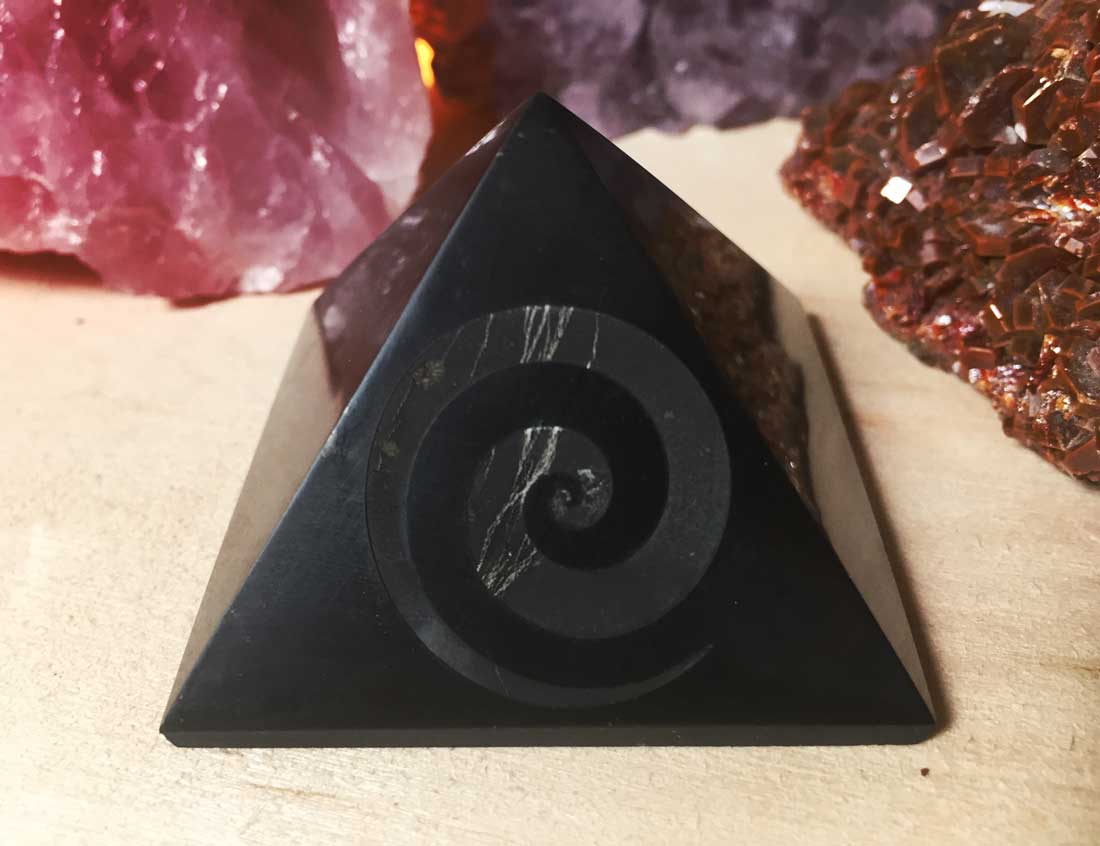 A direct front-facing view of a polished black shungite pyramid with an engraved spiral swirl design on a light wood surface.