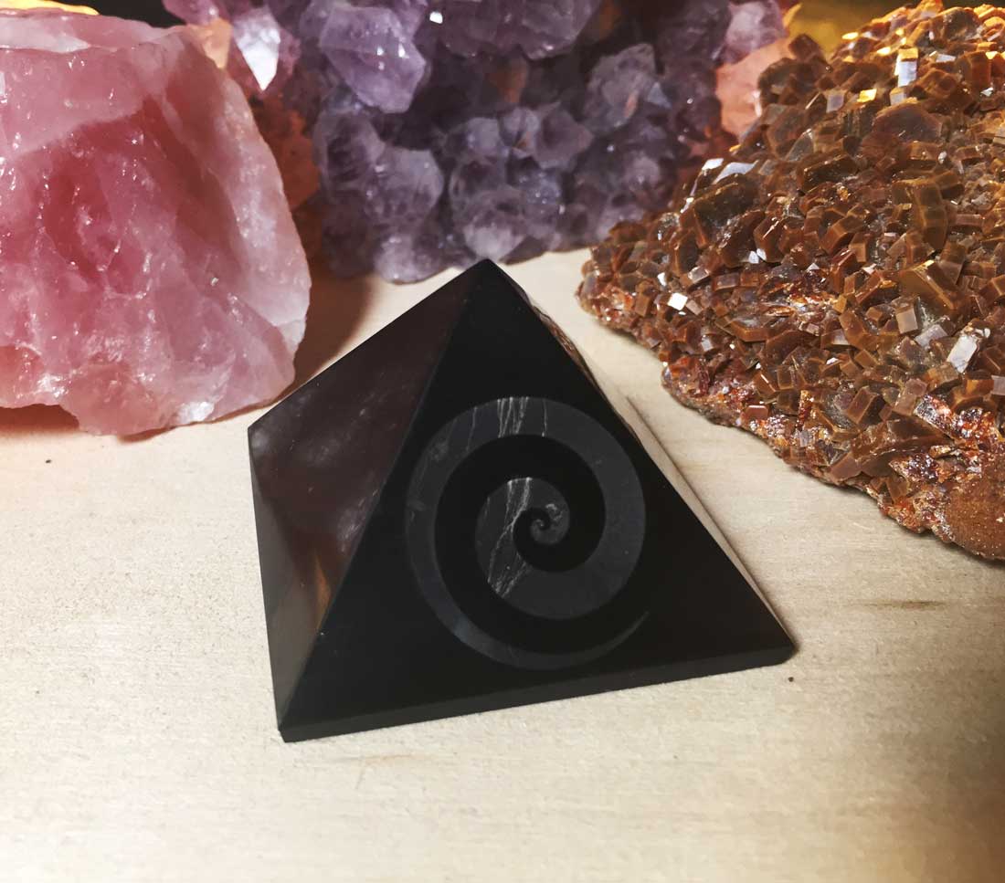 A side-angle perspective view of a polished black shungite pyramid with an engraved spiral swirl design, resting on a wood surface with amethyst clusters.