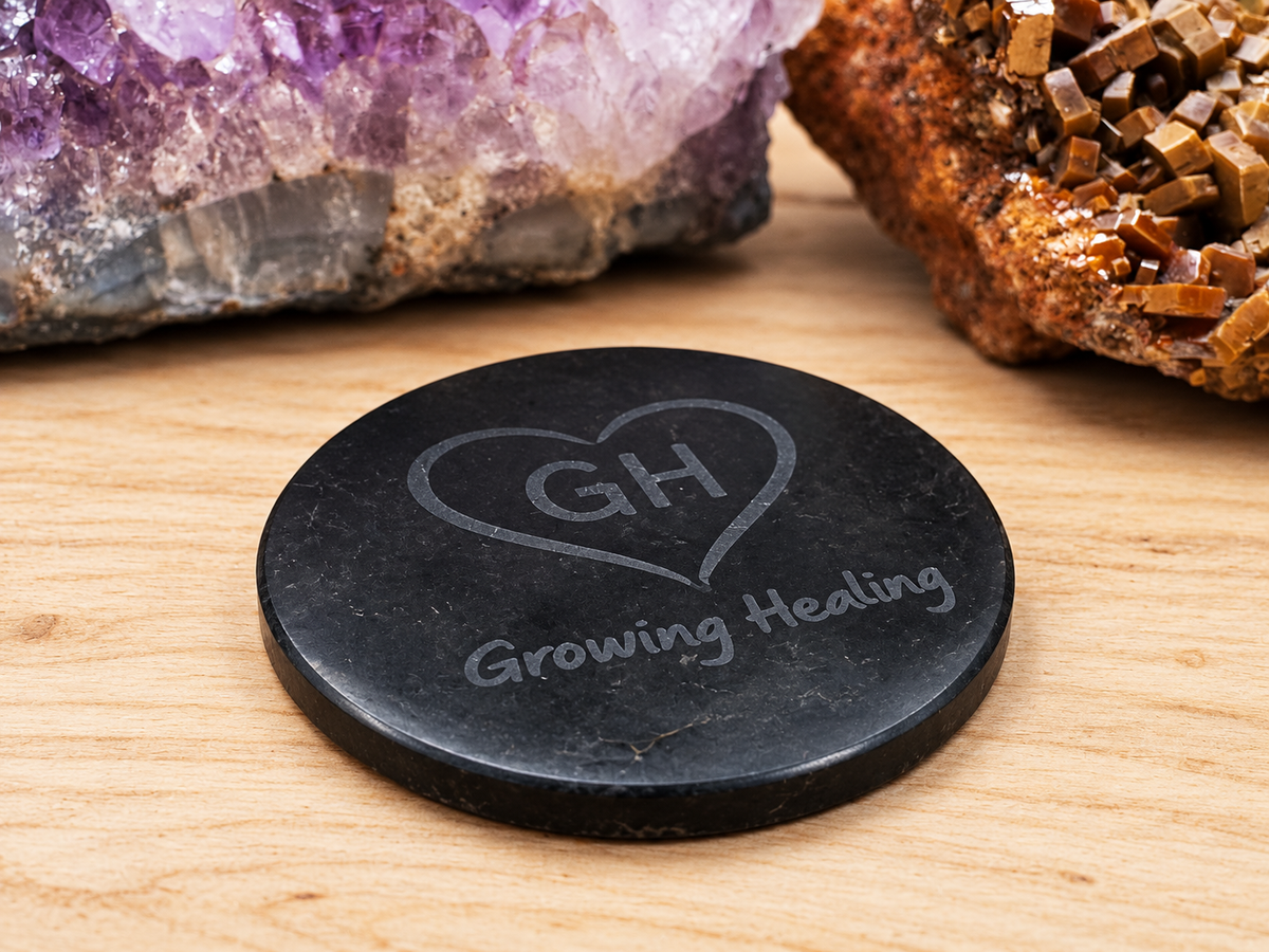 A black circular Shungite tech tab featuring the engraved GH heart logo lying flat on a light wood surface in front of raw purple amethyst and brown vanadinite mineral specimens.