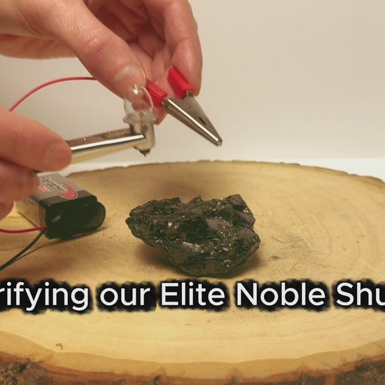 A video demonstration titled Verifying our Elite Noble Shungite, showing hands testing a raw stone with electrical probes and a battery setup on a wooden slab to verify its natural carbon electrical conductivity.
