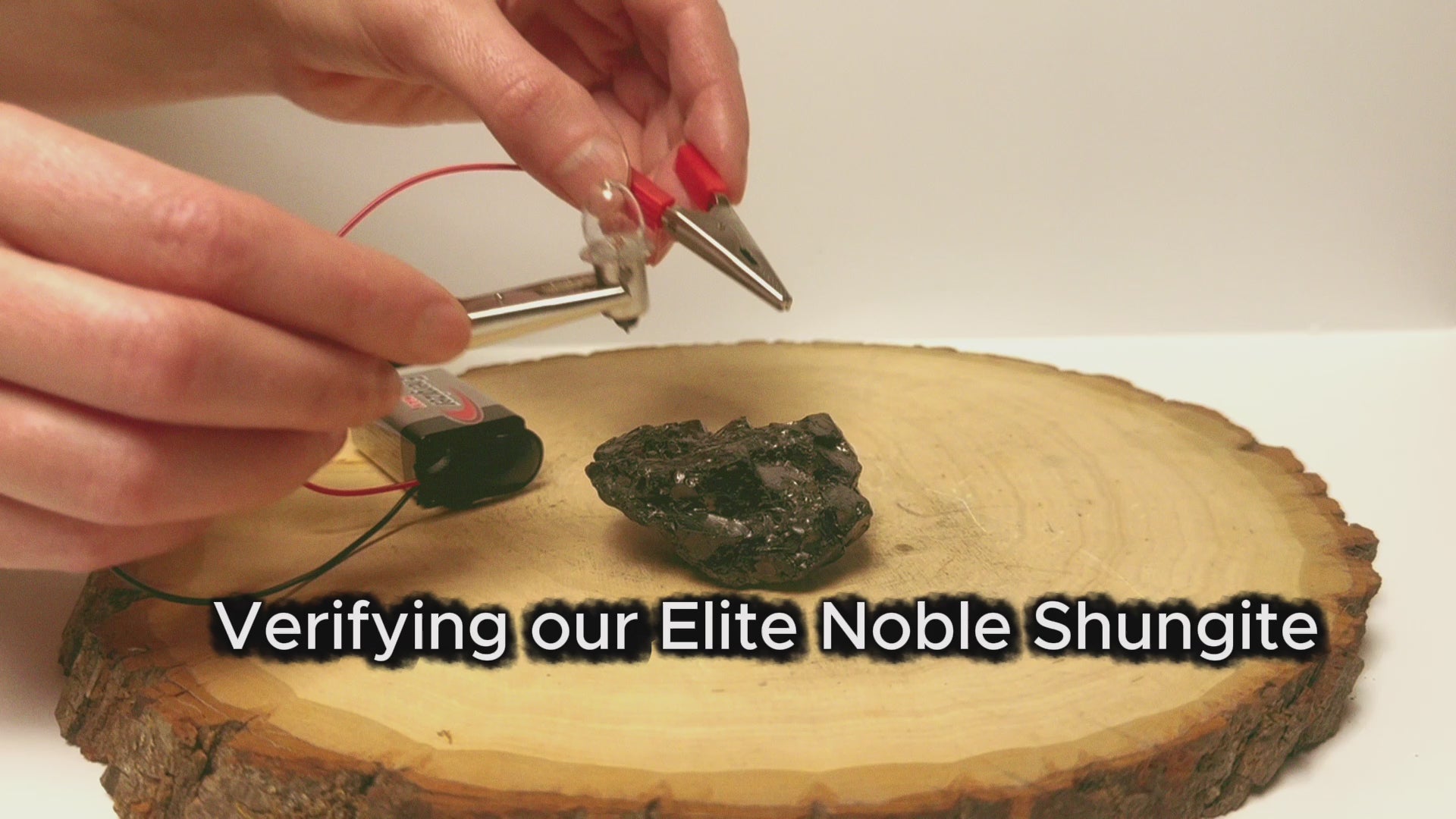 A video demonstration titled Verifying our Elite Noble Shungite, showing hands testing a raw stone with electrical probes and a battery setup on a wooden slab to verify its natural carbon electrical conductivity.