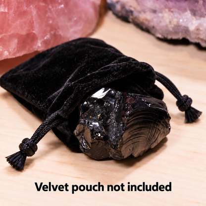 A large, raw elite noble shungite crystal with a metallic silver sheen resting in a black velvet drawstring bag on a wooden base with overlay text reading 'Velvet pouch not included'