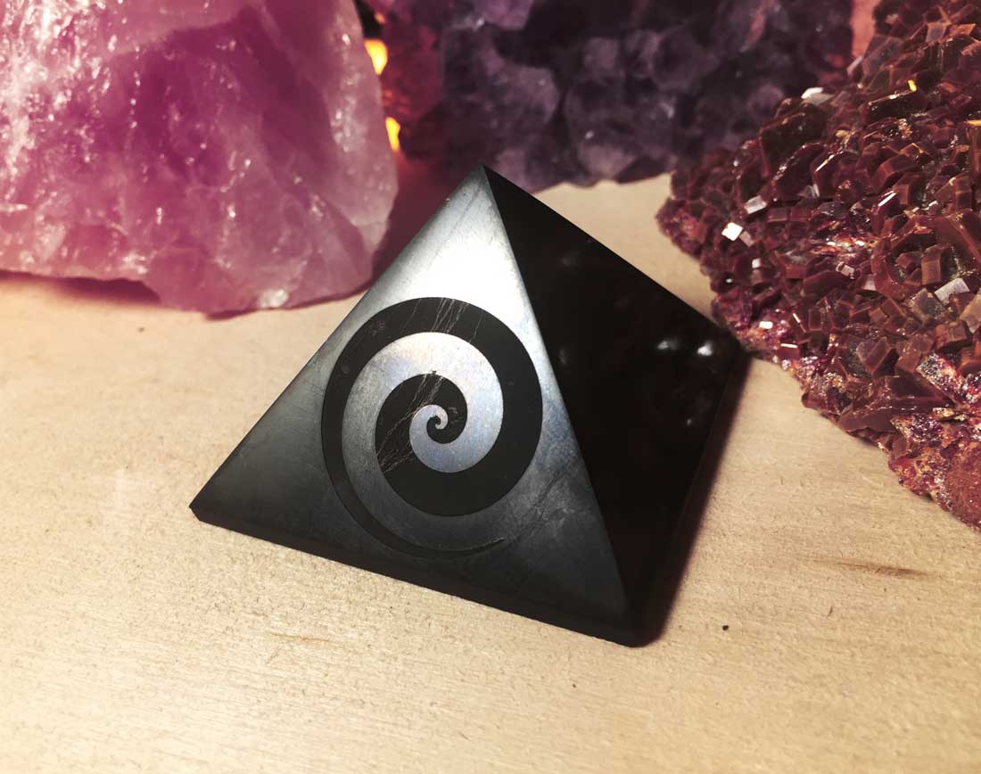 A polished black shungite pyramid engraved with a spiral swirl design, displayed next to crystals.