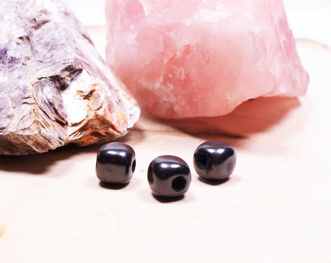 An angled, close-up shot of three loose black Shungite cube beads spread out on a wooden surface, highlighting their drilled holes and metallic luster.
