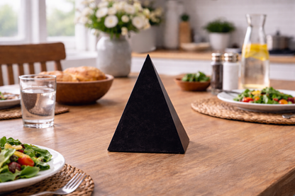 Polished black Shungite Golod Hi-Pyramid displayed on a wooden dining table set with salads, water glasses, and flowers.