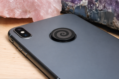 Close up view of a circular shungite phone tab featuring a matte black engraved swirl pattern, displayed on a clean metallic panel surface next to raw pink and purple mineral clusters.