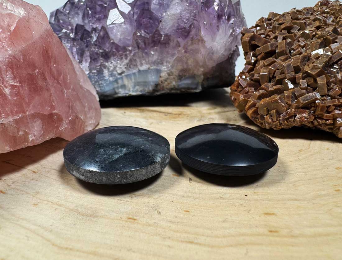 A top-down alternate view of a pair of smooth, circular polished black shungite pocket polarity harmonizer stones resting on a rustic wood slice.
