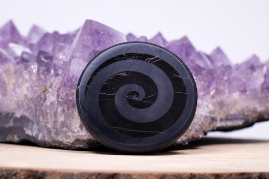 Polished black Shungite pocket stone featuring a hand-carved spiral swirl pattern, leaning upright against a large purple amethyst crystal cluster background.