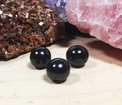 Three highly polished black round Shungite beads arranged in a triangle on a light wood surface, catching reflections of light.