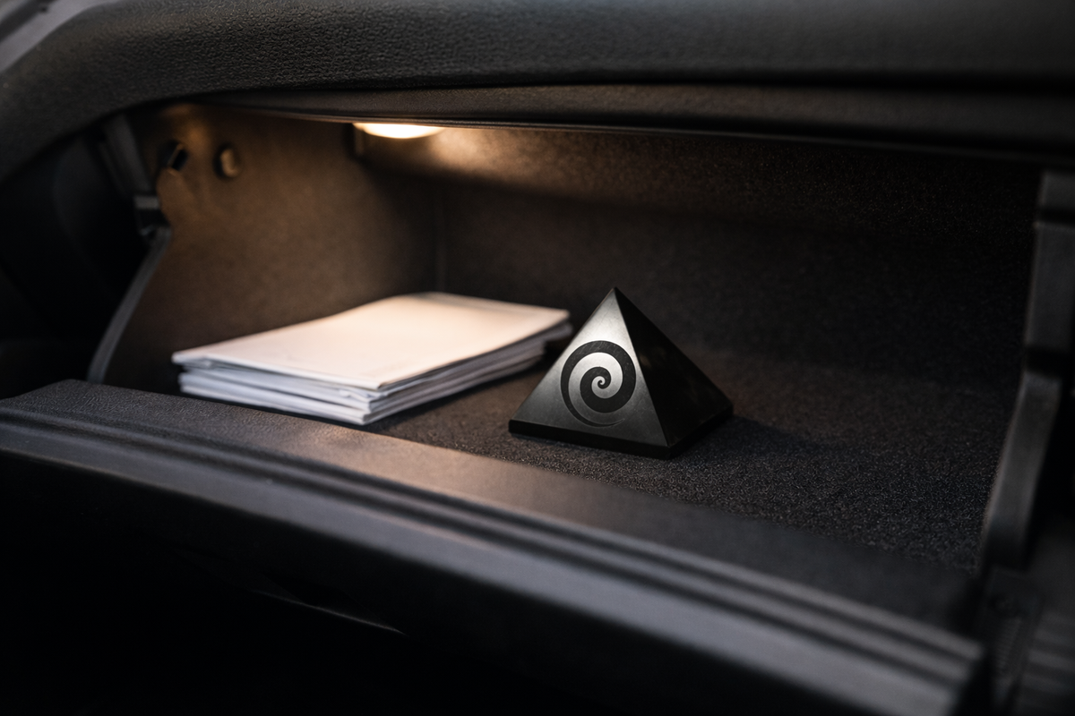 A polished black shungite pyramid with an engraved spiral swirl design sitting inside an open car glove compartment under the interior light.