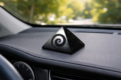 A polished black shungite pyramid with an engraved spiral swirl symbol displayed on a textured car dashboard.