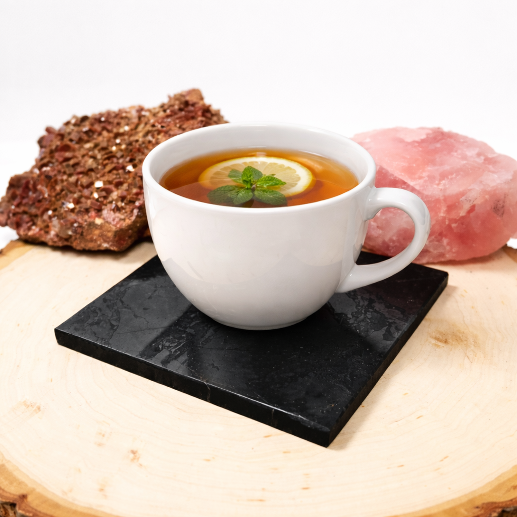 A white ceramic mug filled with tea, lemon, and mint sitting on a square black shungite tile coaster, displayed on a wooden board next to raw quartz crystal clusters.