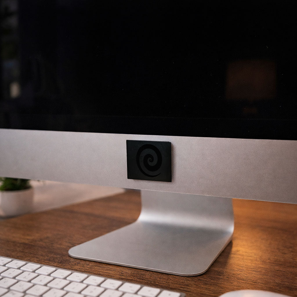 Close up view of a small square black accessory with a carved swirl pattern attached to a brushed aluminum stand panel on a wooden desktop surface.