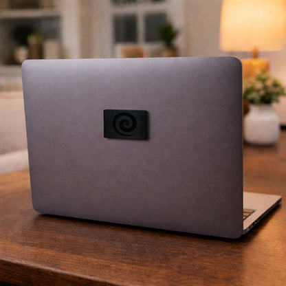 Close up view of a small square black accessory with a carved swirl pattern attached to a space gray matte metallic surface on a wooden tabletop.