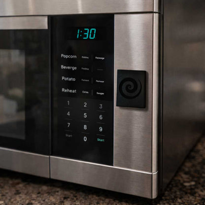 Close up view of a small square black accessory with a carved swirl pattern attached to a brushed stainless steel appliance panel.