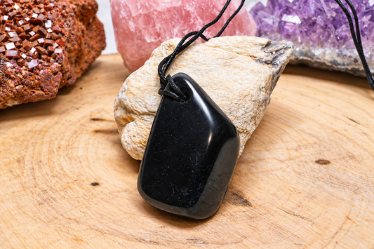 A polished, glossy black Shungite Harmony Pendant attached to a black cord, resting on a textured light beige stone. The background features raw mineral clusters, including brown vanadinite, pink rose quartz, and purple amethyst, arranged on a wooden surface.
