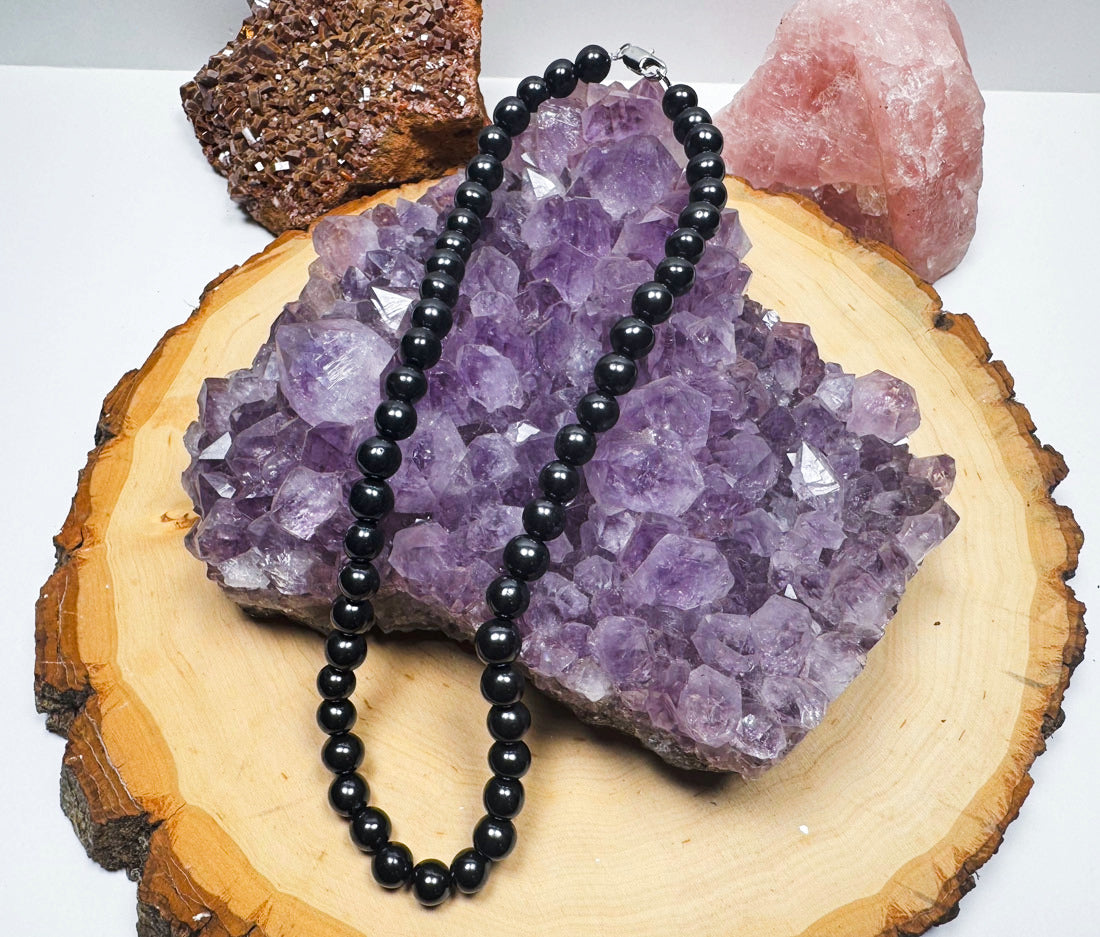 A high-quality strand of glossy black Shungite round beads with a silver clasp, draped elegantly on an amethyst cluster background.