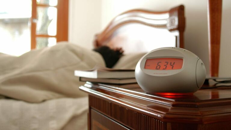 Person in bed sleeping next to a nightstand with an electric alarm clock on it.